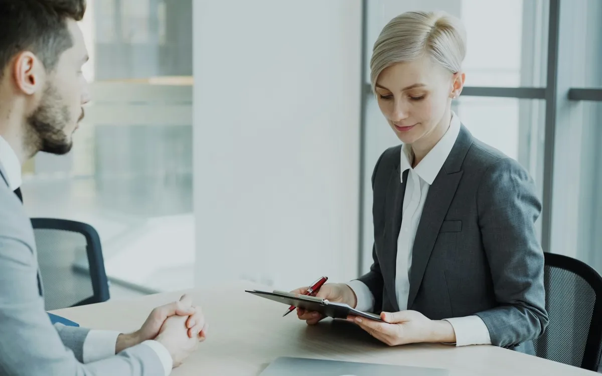 An interviewer making notes on a clipboard across the table from a candidate